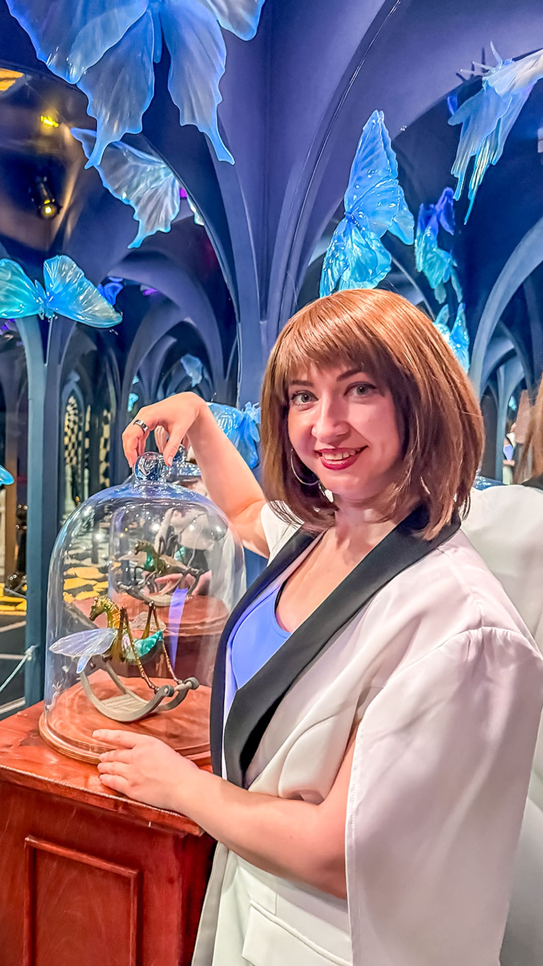 Visitor lifting a glass dome beside glowing blue butterflies in a hall of mirrored arches at Alice in Wonderland Barcelona