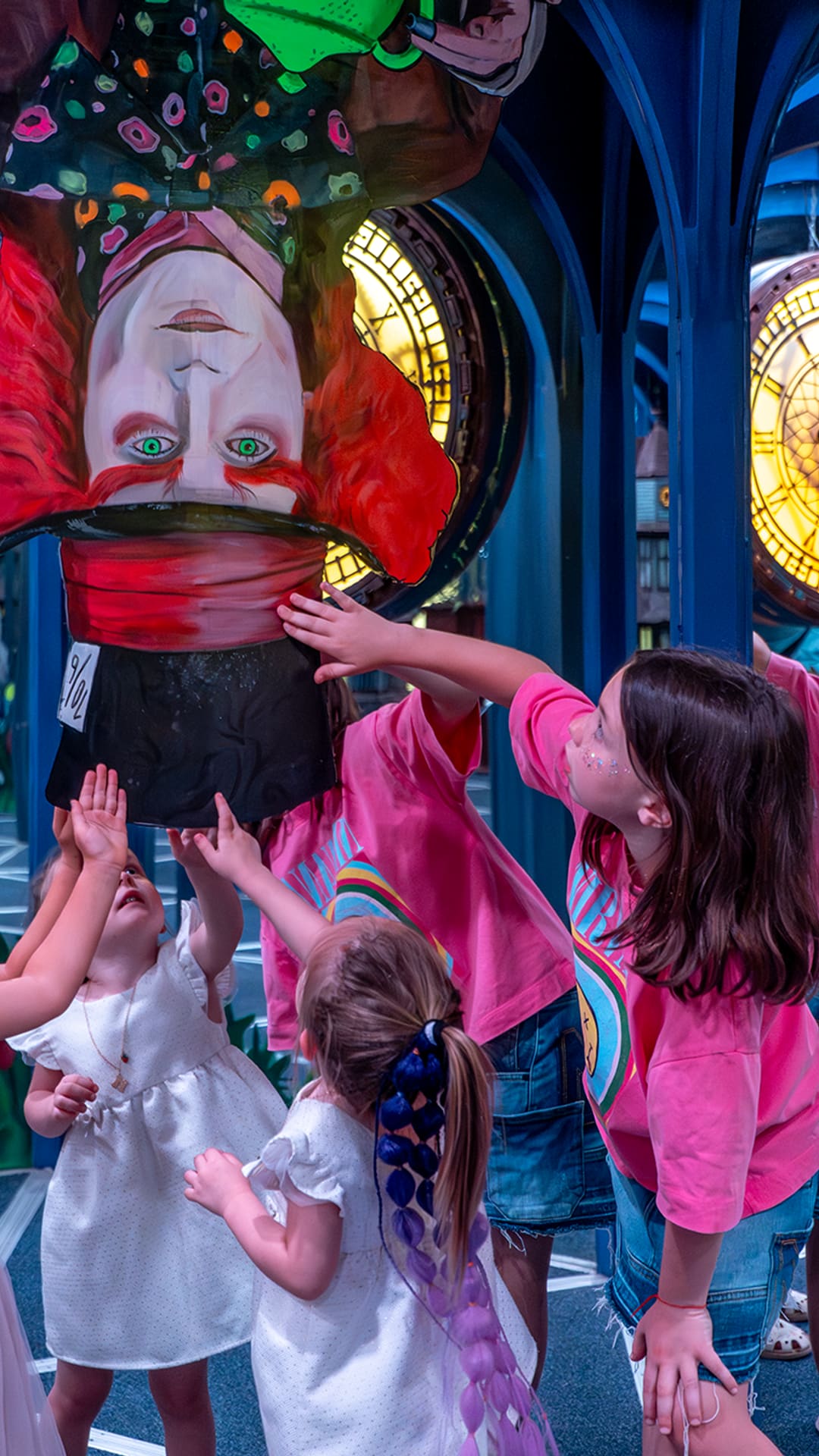 Children reaching up toward an upside-down Mad Hatter figure among glowing clock faces in a mirror room at Alice in Wonderland Barcelona