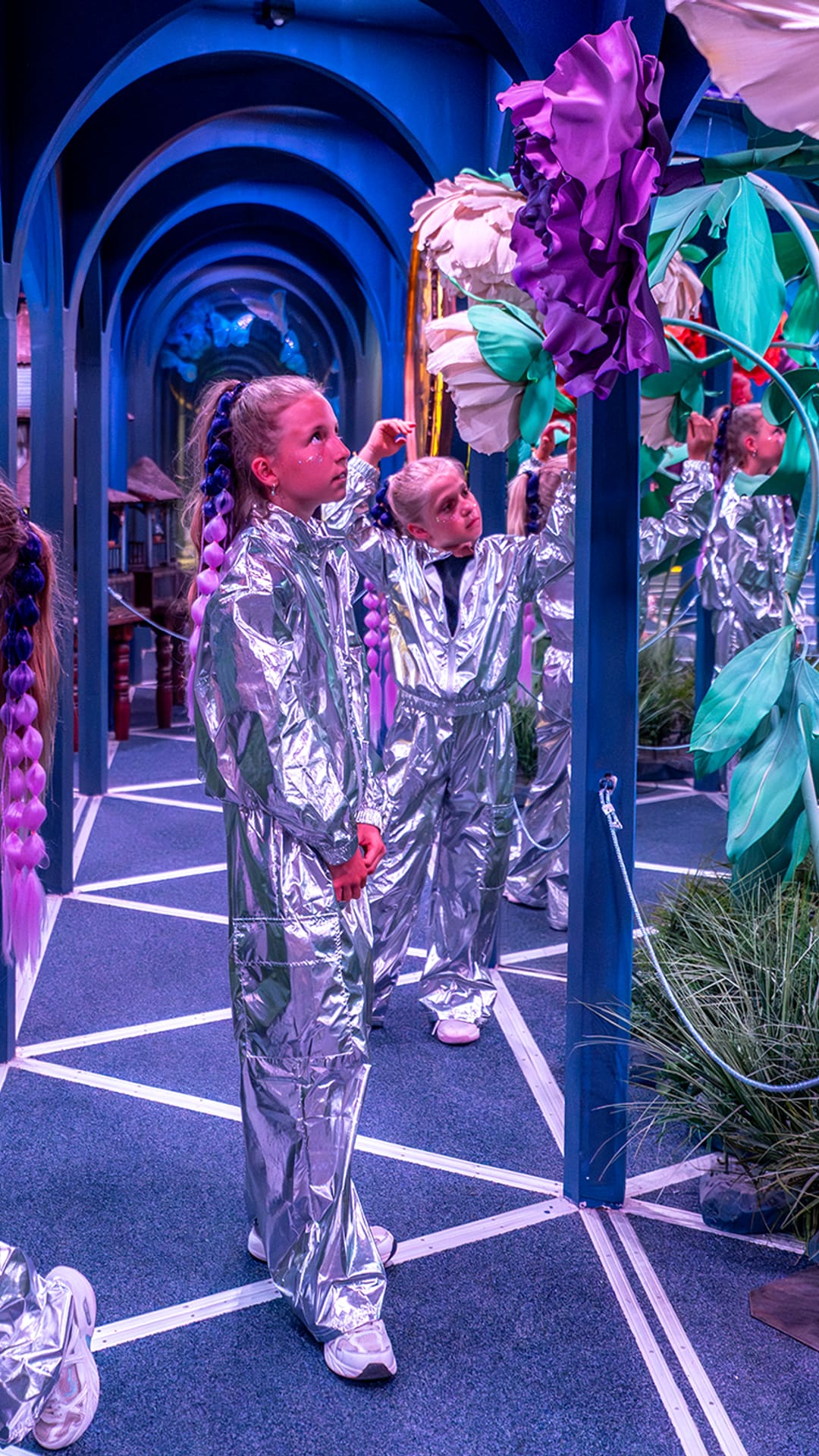 Children in silver outfits reaching toward oversized red and purple paper flowers in the mirrored rose garden at Alice in Wonderland Barcelona